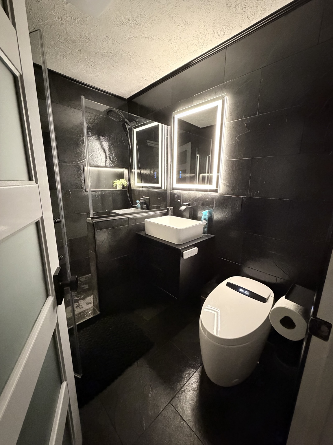 Finished bathroom with black tile, glass shower enclosure, illuminated mirror, and modern fixtures
